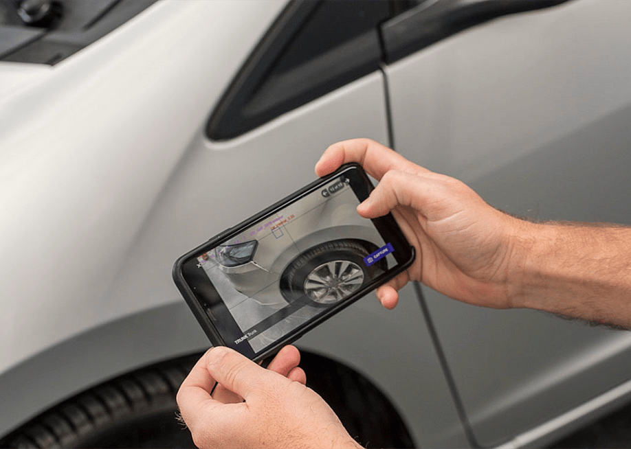 Two hands holding a smart phone aimed at a silver car, the phone's screen shows an AI-powered scan of possible vehicle damage. — Courtesy of Ravin