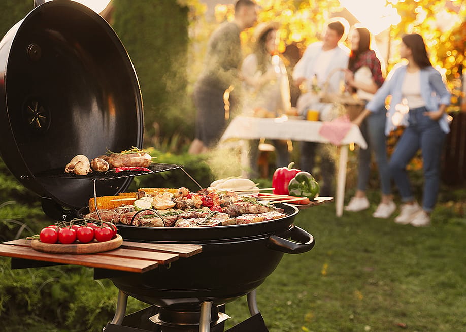 A focus on a barbecue grill set outdoors, with a group of friends having a party in the backdrop. — New Africa // Shutterstock