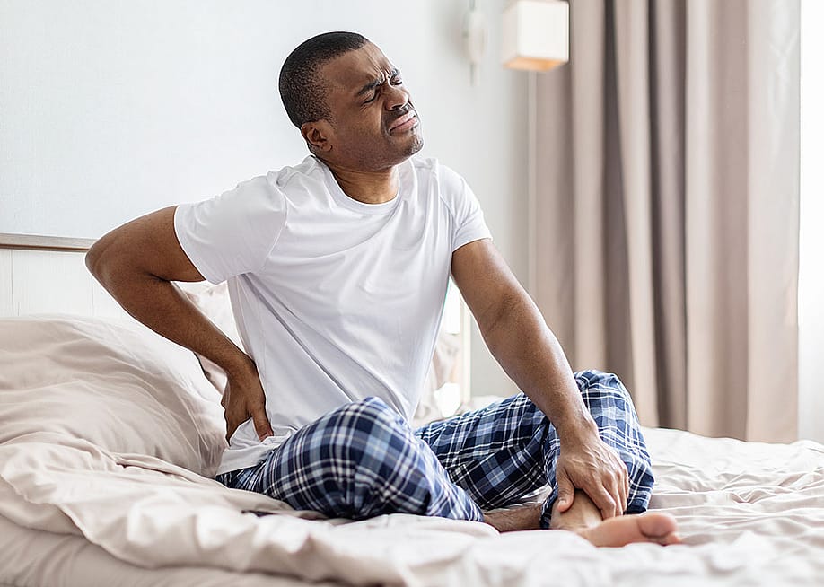 A man sitting on a bed experiencing lower back pain. — FaceStock // Shutterstock
