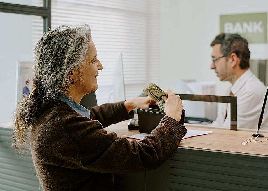 A senior woman handing money to a bank teller. — AnnaStills // Shutterstock