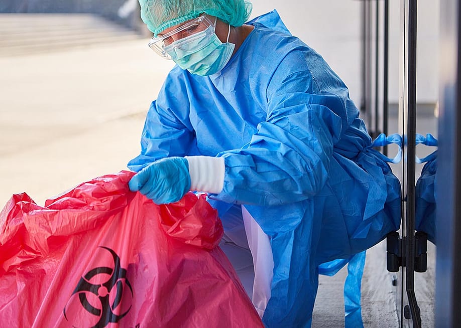 A cleaning staff in full safety gear carefully disposes biohazard waste in a clinic. — Robert Kneschke // Shutterstock
