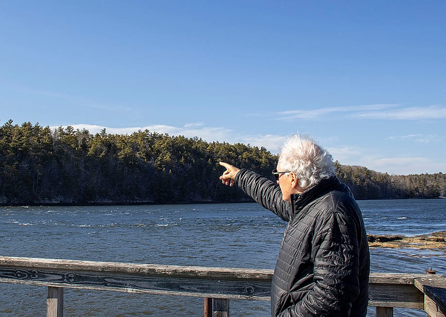 Protect Wiscasset co-founder Richard Davis facing away from the camera wearing a black jacket and his white air blown by the wind, points to the undeveloped land across a waterway where the prospect of a $5 billion data center emerged. — Julia Tilton // The Daily Yonder
