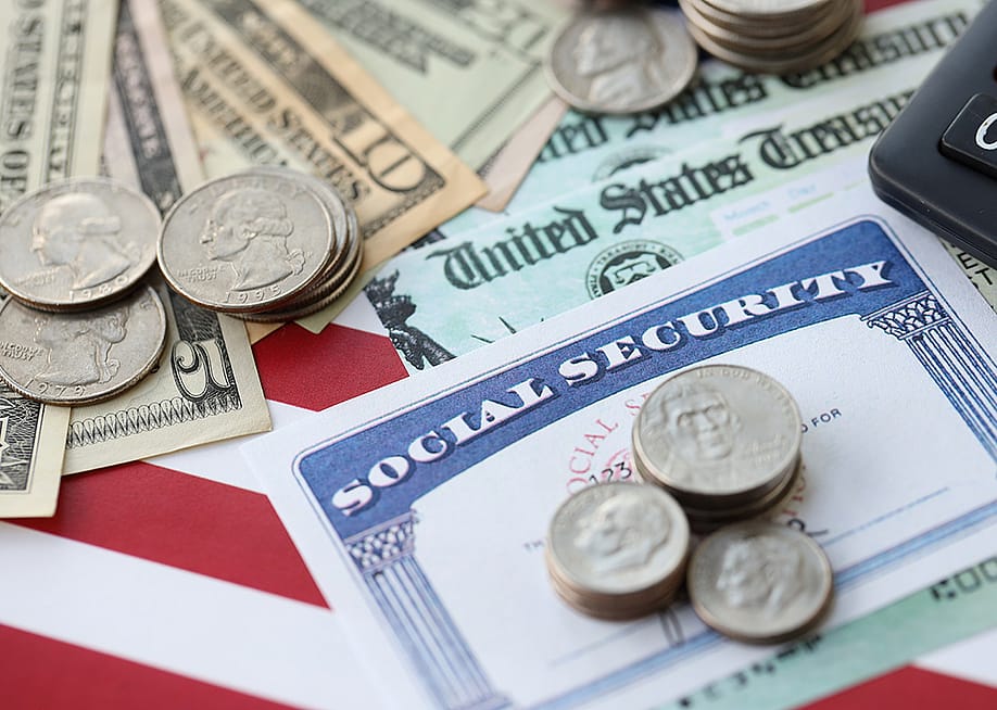 US dollar banknotes and coins, a social security form, and calculator on top of a US flag. — Mehaniq // Shutterstock