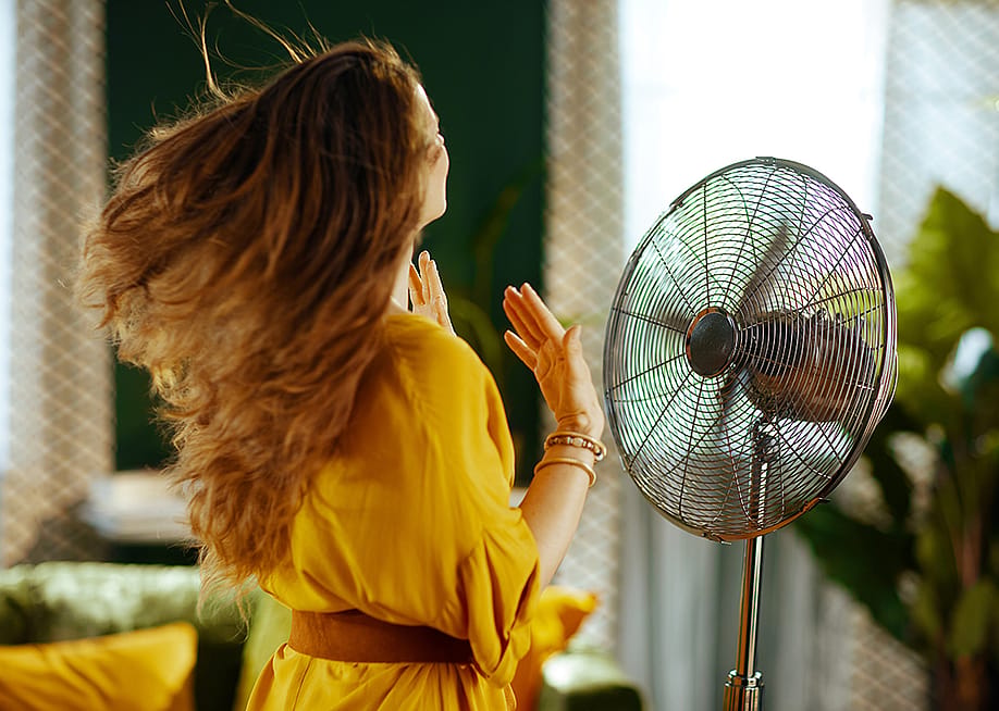 A woman with long hair enjoying the breeze from an electric standing fan. — Alliance Images // Shutterstock
