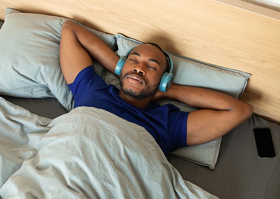 Man lying in bed with his hands on his head and wearing headphones. — Prostock-studio // Shutterstock