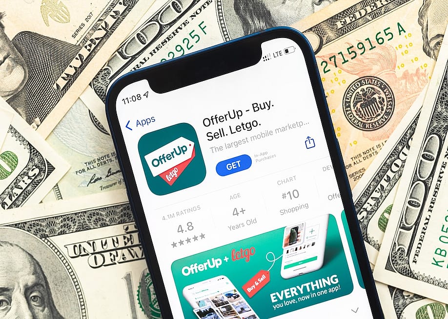 An OfferUP shopping app logo displayed from a mobile phone screen with U.S. dollar bills in the background. — FellowNeko // Shutterstock