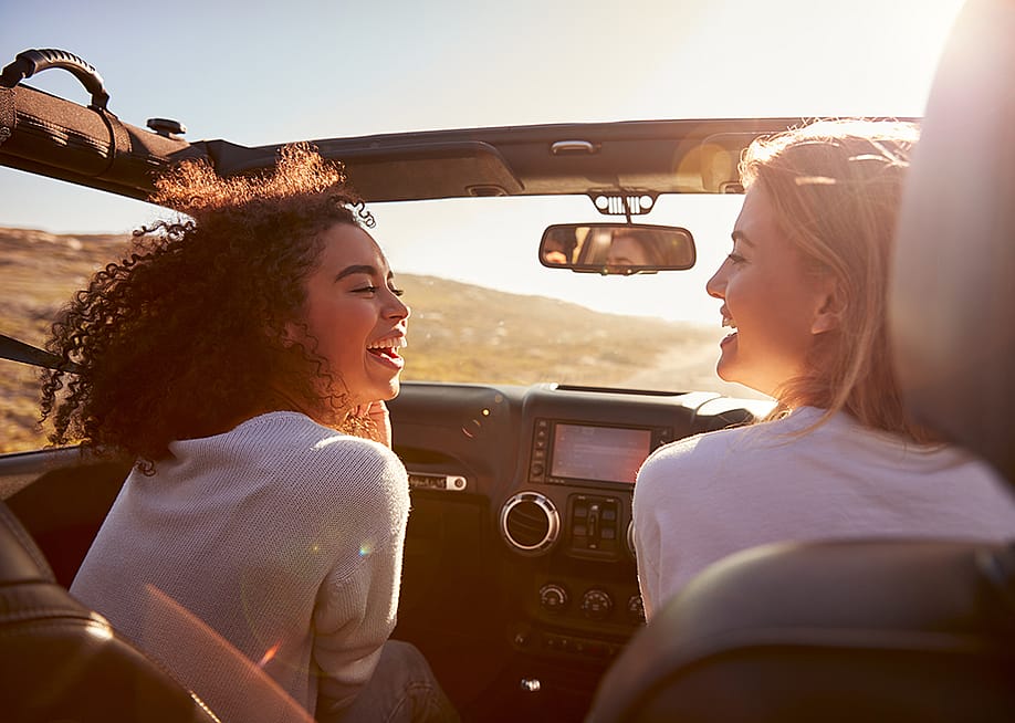 Two female friends happily riding on a highway in their convertible car. — Monkey Business Images // Shutterstock