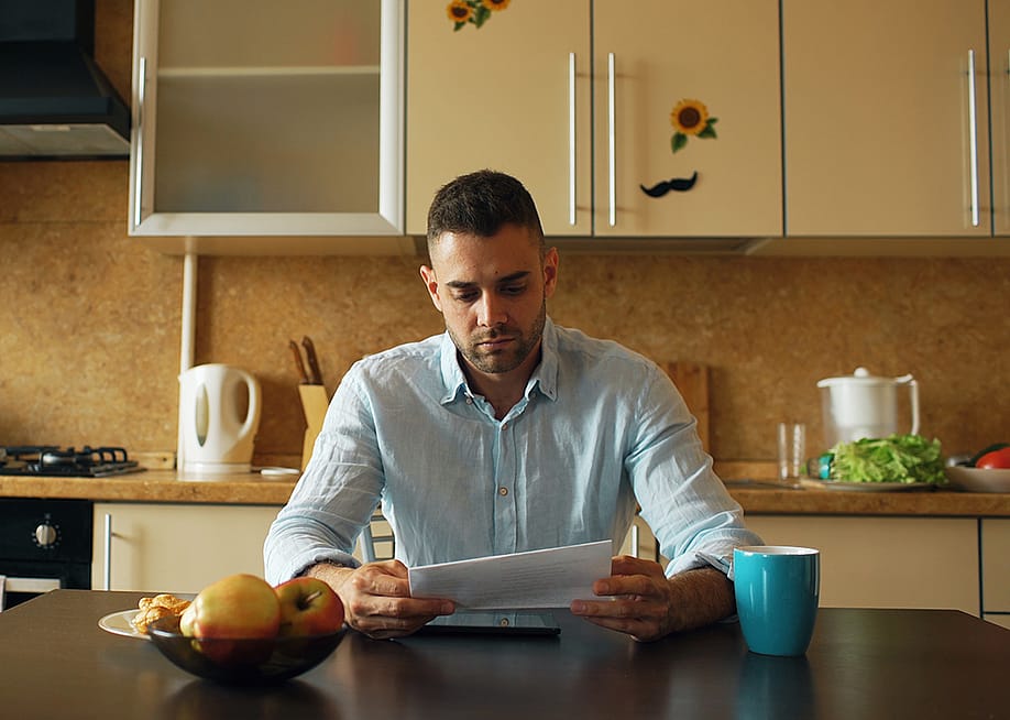 A male homeowner reading an unpaid bill letter in his kitchen. — silverkblackstock // Shutterstock