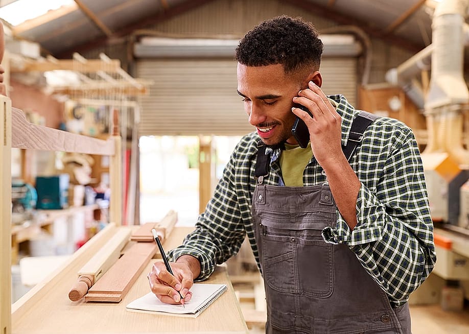 A carpenter taking a customer's order inquiry over a phone call in a furniture work shop. — Monkey Business Images // Shutterstock
