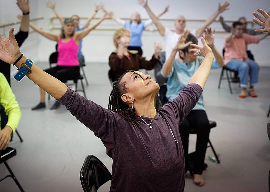 View of upturned face of Black woman with her arms extended, leading a chair dance class with lots of seniors sitting in the background extending their arms in the same motion. — Amber Star Merkens // Dance for PD