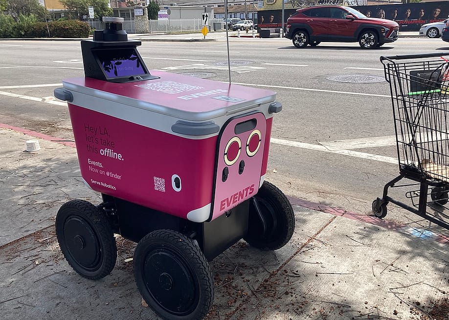 A pink sidewalk delivery robot built by California-based Serve Robotics features an ad from dating app Tinder, while sitting on a sidewalk next to a shopping cart with cars on the road in the background. — Maylin Tu for Next City