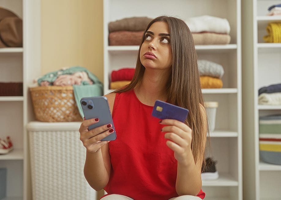 A young adult appearing to be frustrated while holding her smartphone and credit card in a dressing room. — Krakenimages.com // Shutterstock