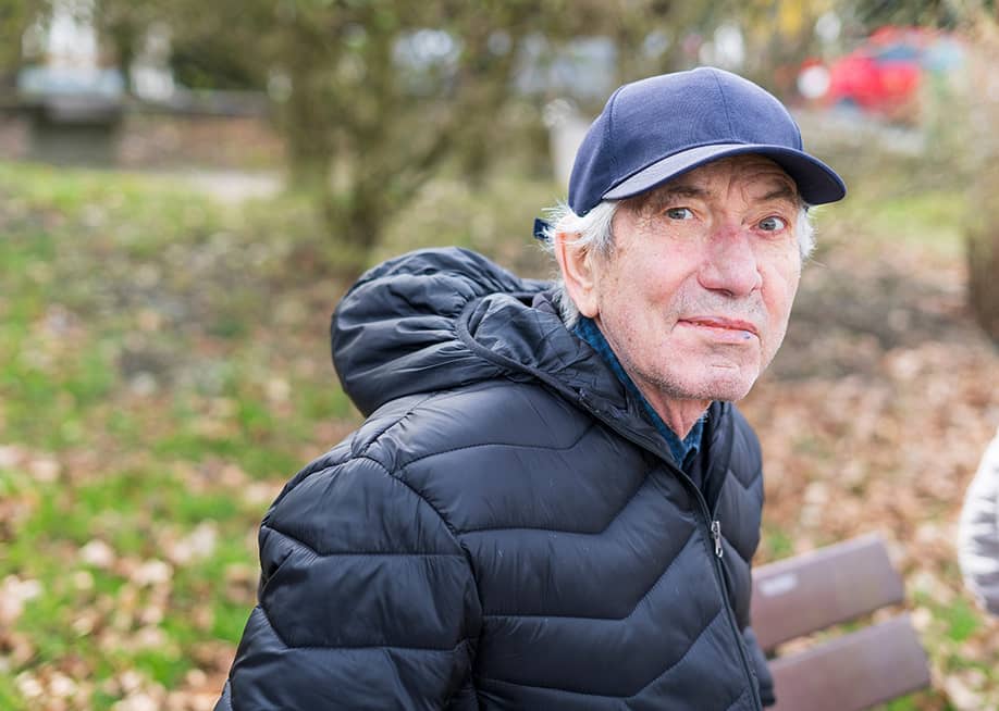 An elderly man wearing a thick jacket and hat while sitting alone on a bench in a park. — PeterPike // Shutterstock