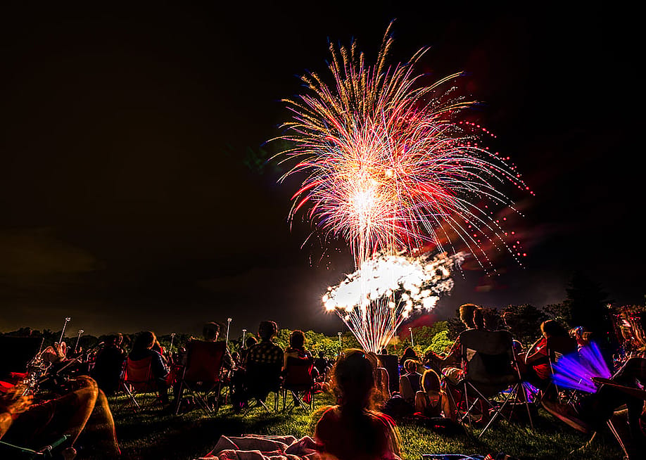Crowds of people sit in lawn chairs and blankets to enjoy the colorful fourth of July independence day celebratory fireworks exploding in the black night sky. — Big Joe // Shutterstock