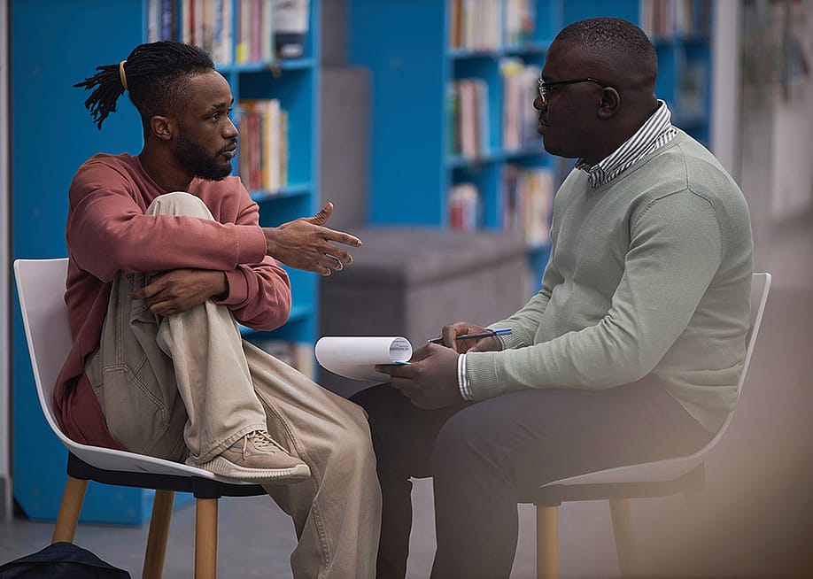 A young black male student talking to a counselor in their college library. — SeventyFour // Shutterstock
