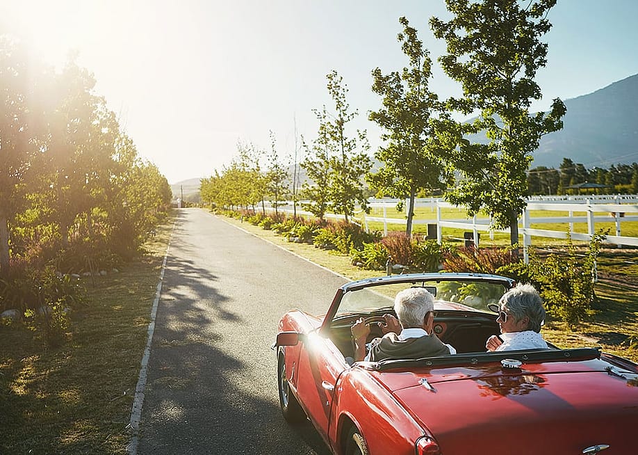 An elderly couple driving in a convertible car for a road trip on a summer day. — PeopleImages // Shutterstock
