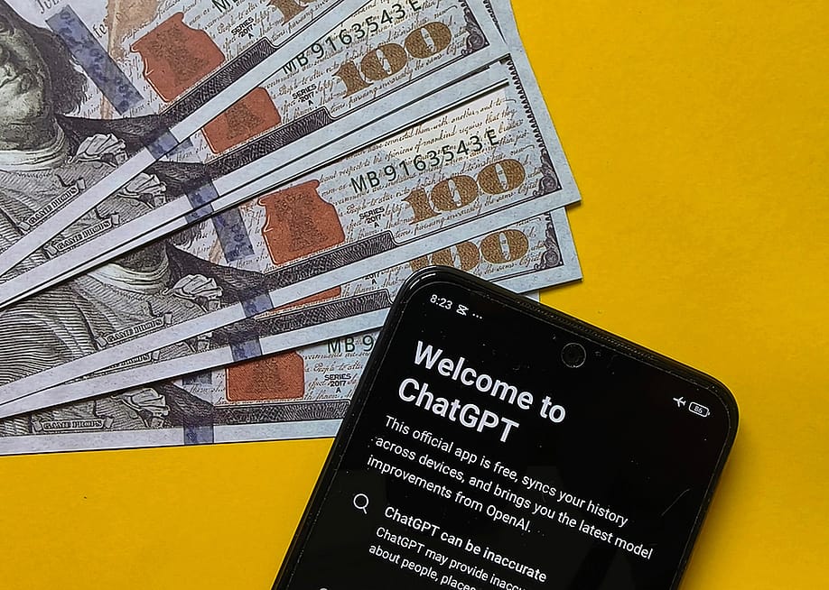 US 100 dollar banknotes and a smartphone displaying ChatGPT in a yellow background, symbolizing the concept of financial AI. — For East // Shutterstock