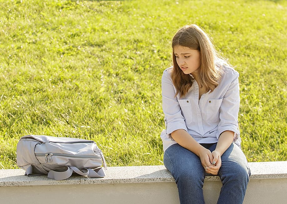 An upset teenage girl sitting on a curb and staring at her backpack for school. — Larysa Dubinska // Shutterstock