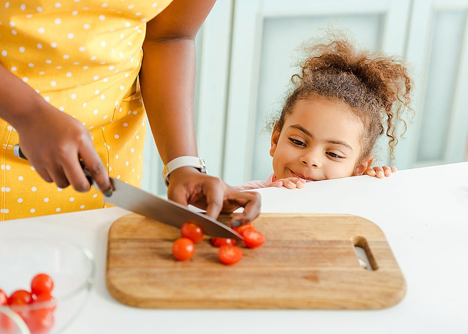 A little child watching her mother slice cherry tomatoes on a chopping board in the kitchen. — LightField Studios // Shutterstock