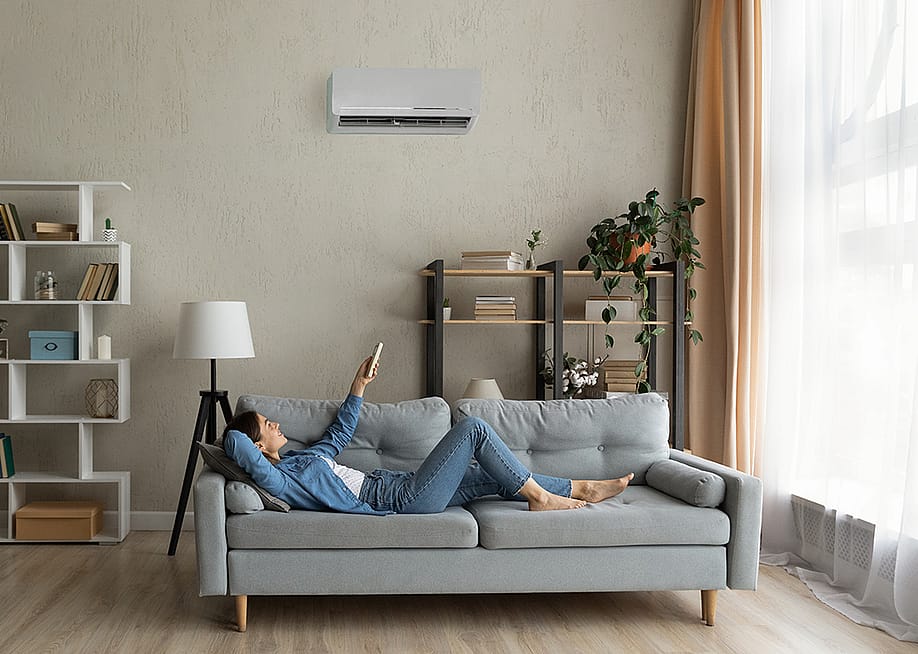 A young woman at home relaxing on the sofa and turning up the AC temperature. — fizkes // Shutterstock