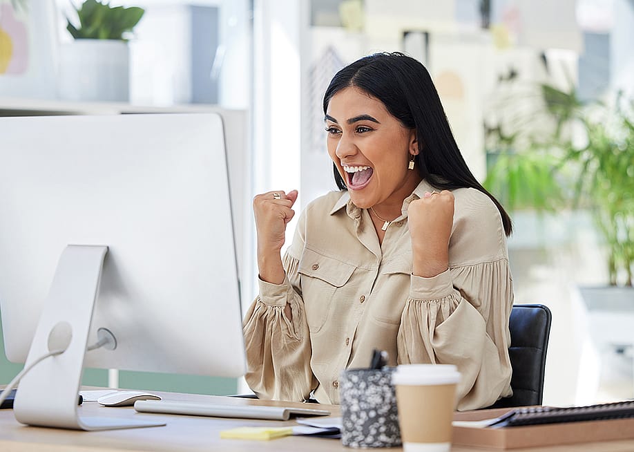 An enthusiastic businesswoman celebrates in front of her computer monitor at work. — PeopleImages // Shutterstock
