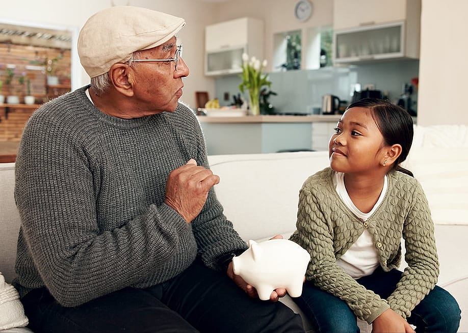 A grandfather teaching his young granddaughter how to save money with a piggy bank. — PeopleImages // Shutterstock
