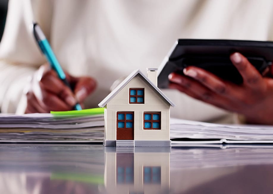 A small figure of a home with a real estate professional calculating finances in the background. — Andrey_Popov // Shutterstock