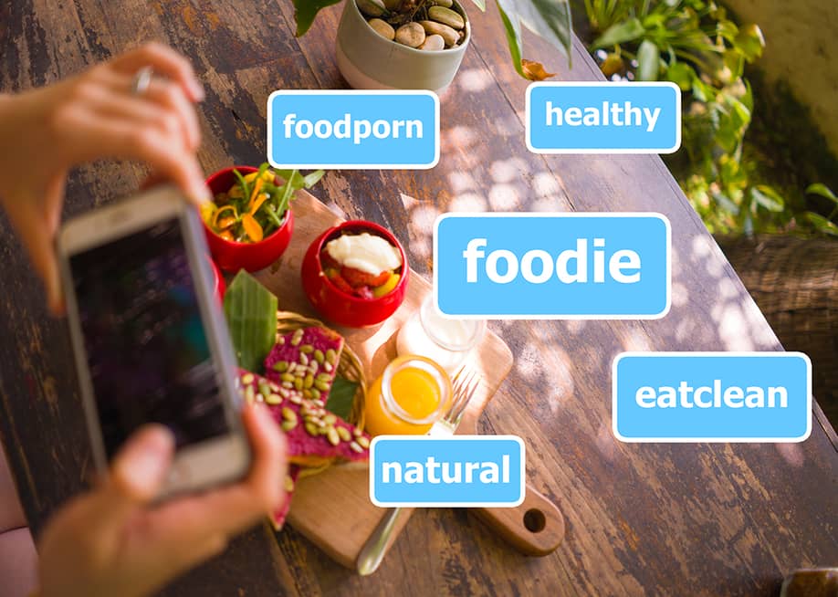 Person taking a photo of their food illustrated with common social media hashtags. — TheVisualsYouNeed // Shutterstock
