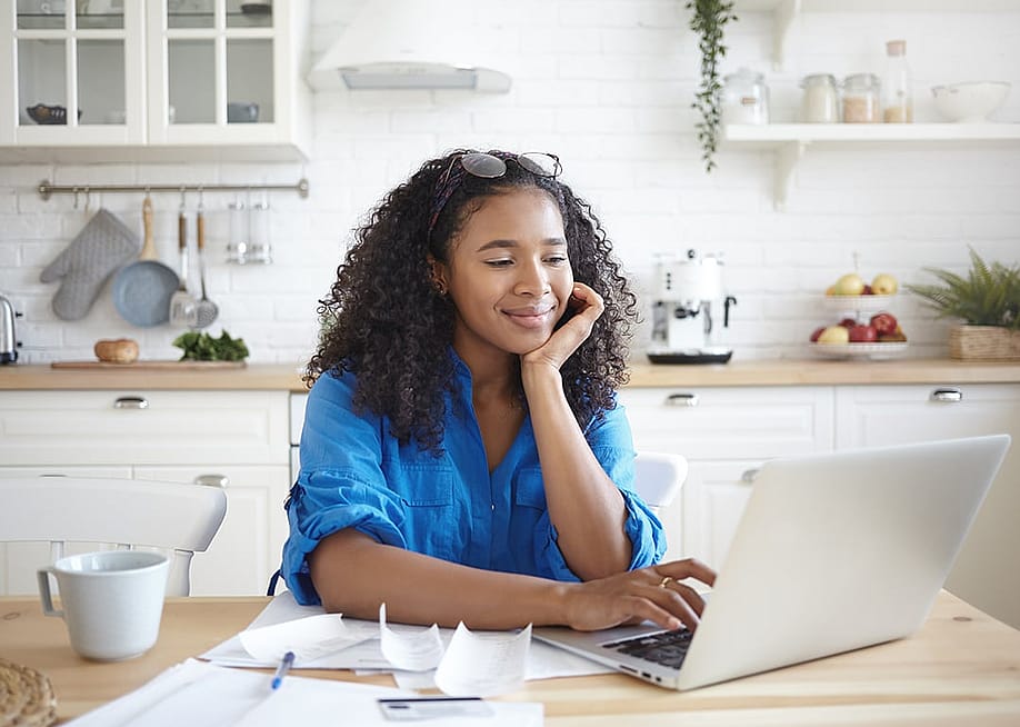 A young woman paying bills online in her home kitchen. — shurkin_son // Shutterstock