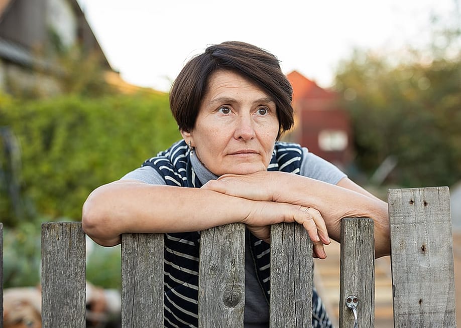 An elderly person leaning on a wooden fence outdoors. — Caftor // Shutterstock