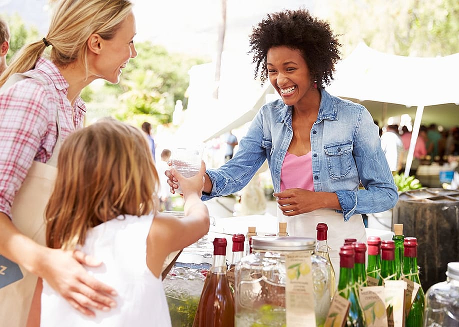A young businesswoman selling drinks to a mother and daughter at a farmer's market stall. — Monkey Business Images // Shutterstock