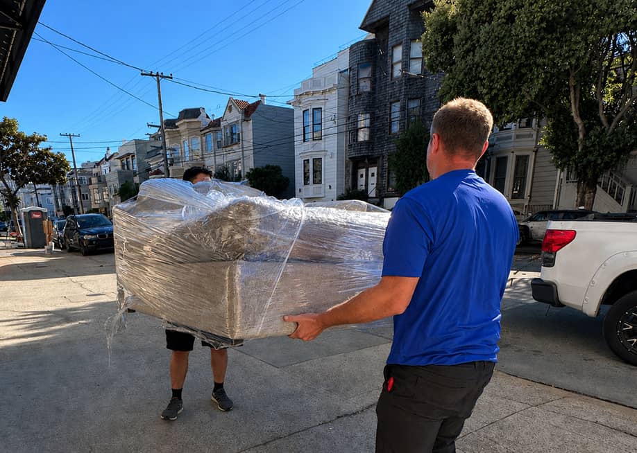 Professional movers carrying a plastic-wrapped couch across a street. — Lugg