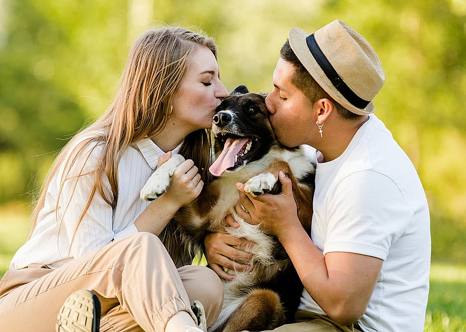 A young couple kissing their pet dog outdoors in a park. — Daniel Megias // Shutterstock