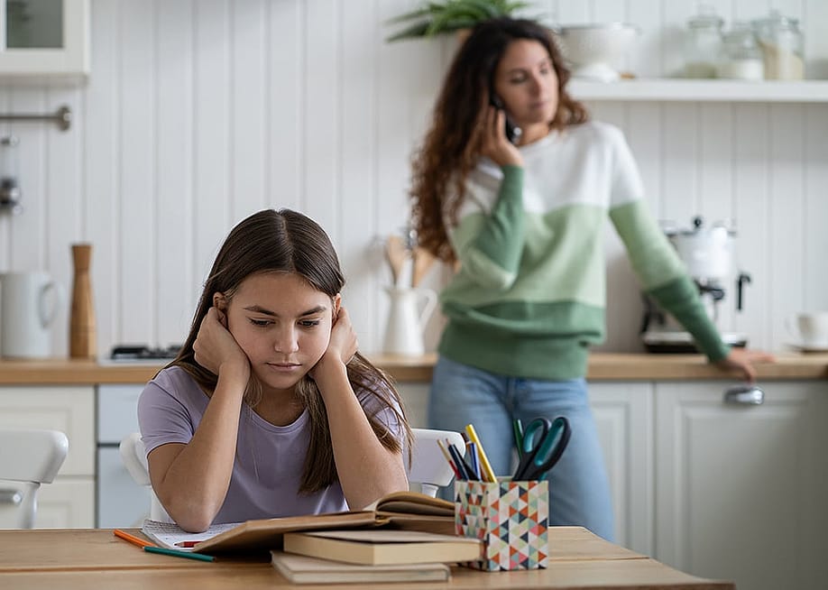 A parent talking to someone over the phone, with her child also struggling to do homework in the dining area. — DimaBerlin // Shutterstock
