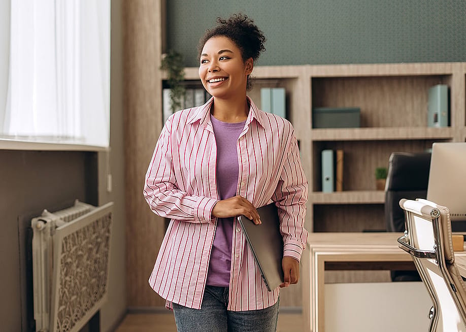 A happy businesswoman in an office walking with her laptop. — AYO Production // Shutterstock