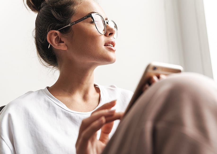 A young adult looking afar out the window after receiving a suspicious business text from her smartphone. — Dean Drobot // Shutterstock