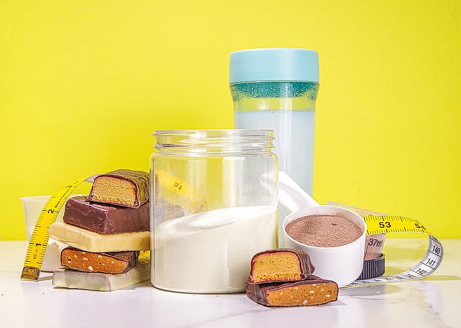 Protein bars, nutrition drink, supplement powder, and measuring tape placed in a table with a yellow background. — Rimma Bondarenko // Shutterstock