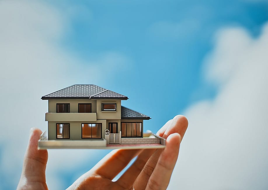 A person's hand holding a miniature house with the bright sky in the background. — beeboys // Shutterstock