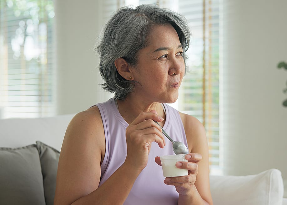 A middle-aged woman at home eating a cup of yogurt. — Chay_Tee // Shutterstock