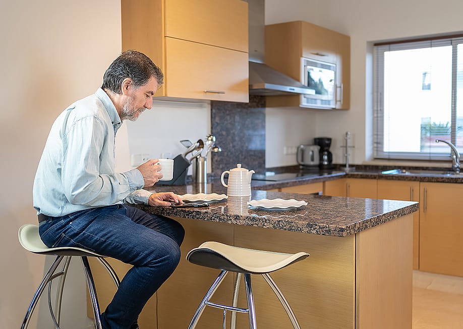 Senior man sitting on a stool in the kitchen, drinking a cup of coffee and looking at the mobile phone. — Jose Miguel Sanchez // Shutterstock