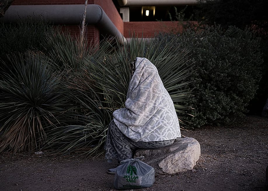 An unhoused person wrapped in blanket and sitting on a rock outdoors in Tucson, Arizona. — ERNESTO BENAVIDES // AFP via Getty Images