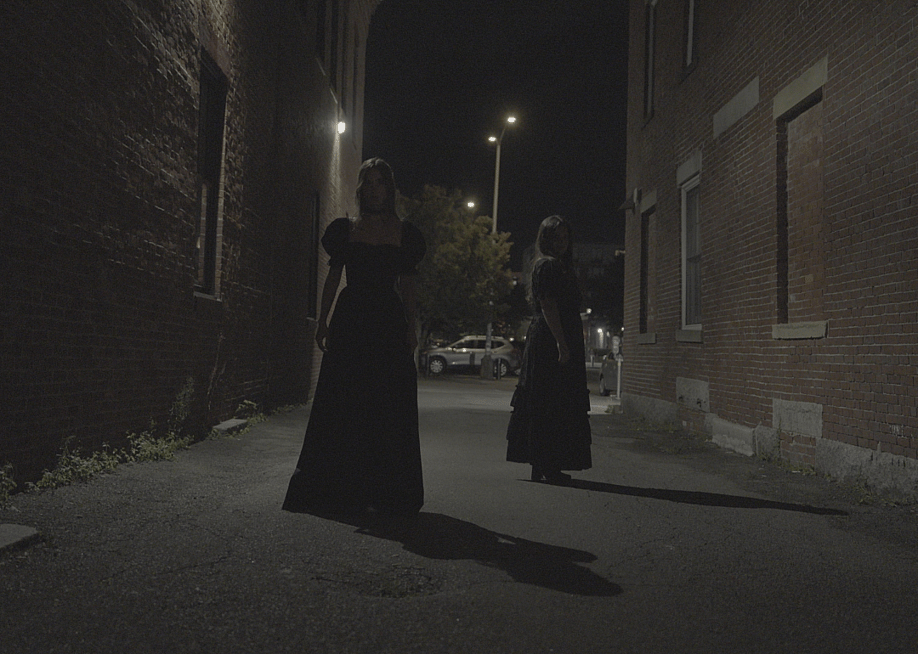 Two figures in black dresses stand in a dark brick alleyway at night. — Sony Music Entertainment