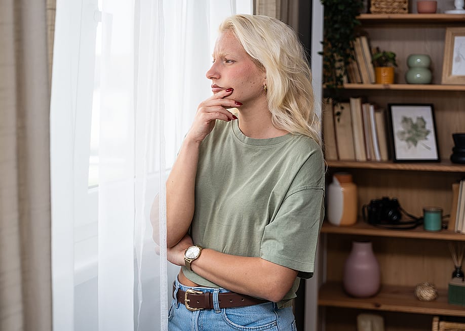 A pensive young woman at home looking through her window. — Srdjan Randjelovic // Shutterstock