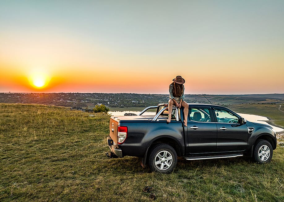 A person sitting on the roof of a pickup truck parked on a grassy hill during sunset. — Sid0601 // Shutterstock