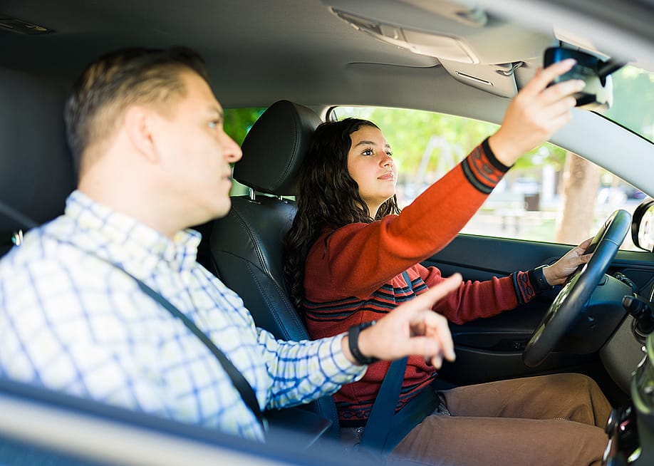 A teenage girl adjusting rearview mirror while learning to drive with her father. — antoniodiaz // Shutterstock
