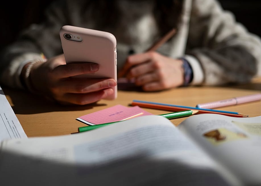 A young student holding a smartphone at a desk with an open book, colored pencils, and sticky notes. — Marijan Murat // picture alliance via Getty Images