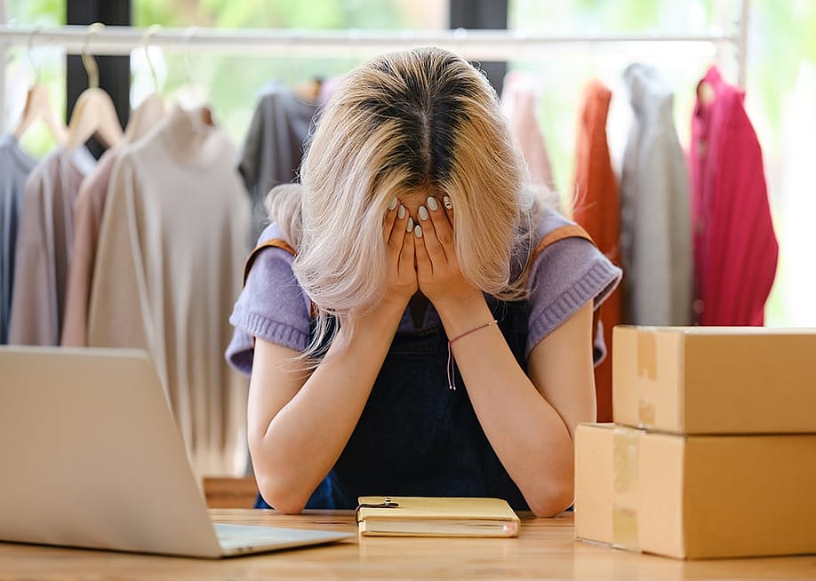 A young female entrepreneur working in her studio while covering her face in apparent distress. — Treerat Wongvorapat // Shutterstock