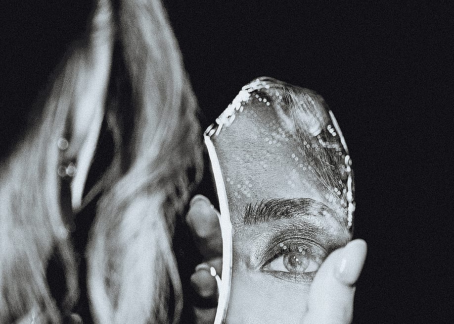 A black-and-white portrait of a woman holding a broken piece of a mirror that reflects a close-up of her eye. — FaceStock // Shutterstock
