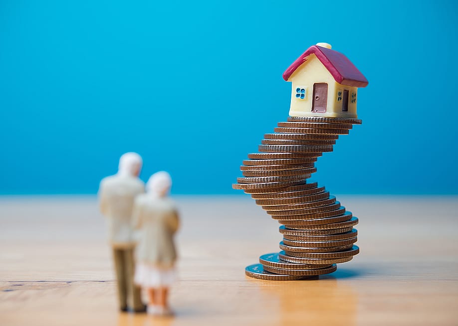 A miniature senior couple standing before a toy house on unstable stack of coins, representing financial risk with longer loans during retirement. — Pla2na // Shutterstock