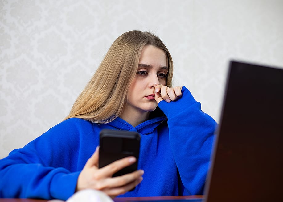A young woman sits frozen in front of her laptop while holding her phone. — Timur Malazoniia // Shutterstock